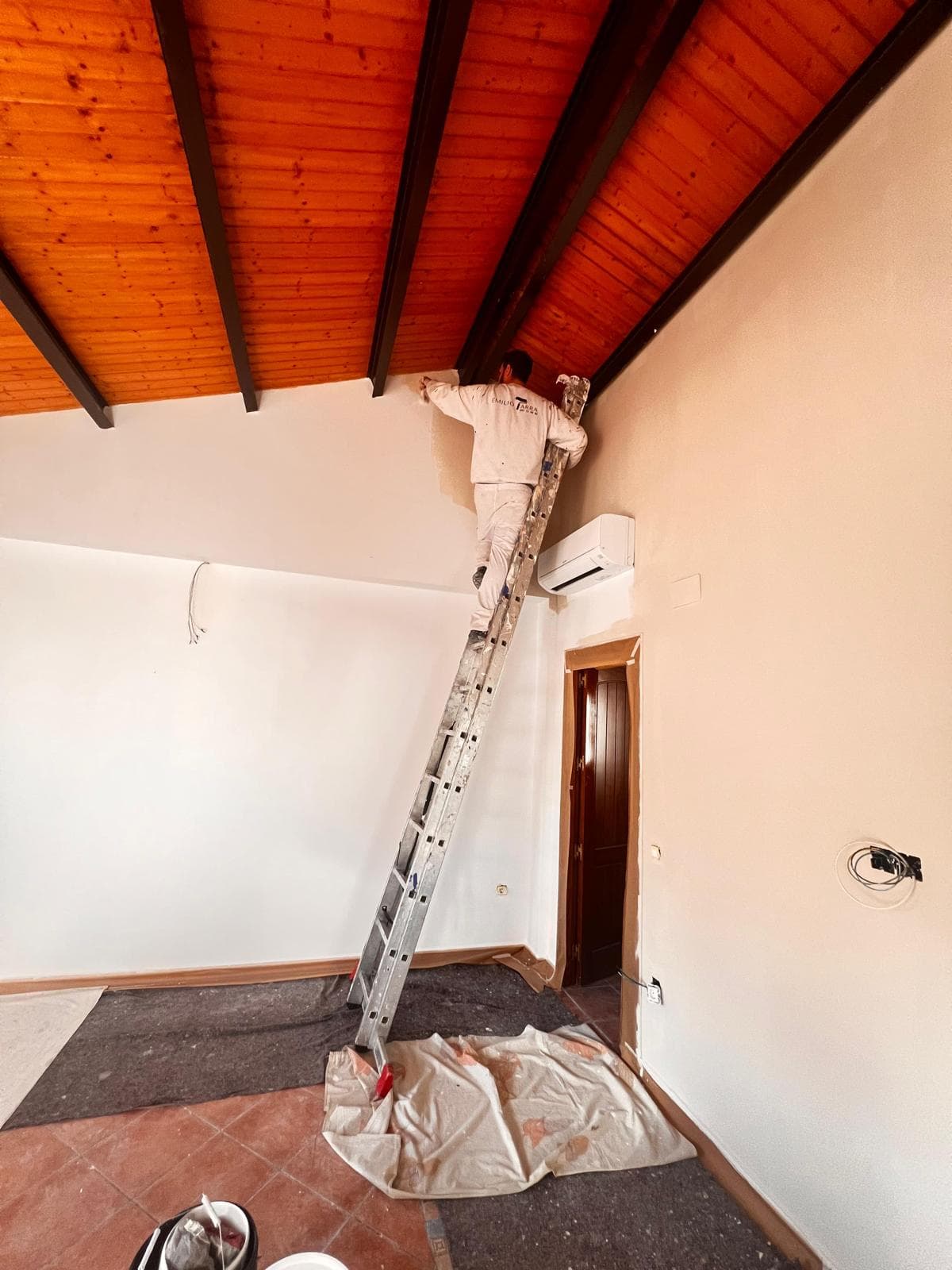 Emilio pintando en lo alto de una escalera, bajo un techo de madera